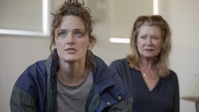 A young woman with tousled hair and a blue jacket sits in the foreground, looking intently ahead. An older woman with blonde hair and a blue blouse sits behind her, watching with a concerned expression.