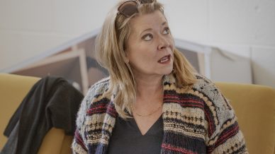 A woman with blonde hair and sunglasses on her head sits on a yellow chair, using a laptop. She wears a striped jumper and looks to her left with a surprised expression. A black jacket is draped over the chair.