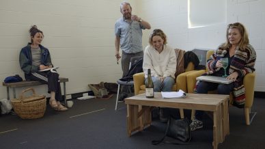 Four adults sit and stand in a casual, bright room with white walls. Three women are seated, smiling, with notebooks. A man stands behind them, holding a drink. A bottle and glasses are on a wooden coffee table in front.