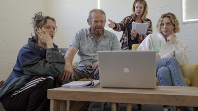 Four people sit around a wooden coffee table, looking intently at a laptop. One person is pointing at the screen, while others appear focused or thoughtful. Papers and a glass of wine are on the table.