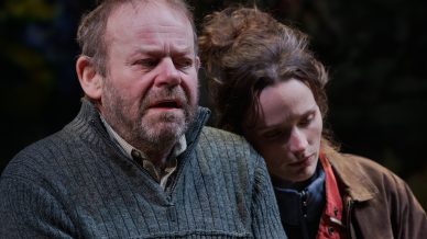 A bearded man in a grey jumper looks upset, with watery eyes, while a woman with brown hair rests her head gently on his shoulder, comforting him. Both appear emotional, set against a dark, blurred background.