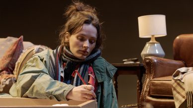 A woman in a green jacket sits on the floor next to a cardboard box, looking down thoughtfully while holding a pencil. Behind her are a lamp, armchair, and a side table with scattered items.