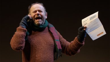 A man wearing a brown jumper, striped scarf, and black gloves speaks on a phone whilst holding a piece of paper, smiling with an expressive face against a dark background.