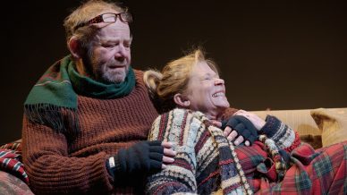 An older man and woman sit closely on a sofa, bundled in warm jumpers, scarves, and gloves. The man looks content while the woman leans back, smiling and laughing, creating a warm and cosy atmosphere.