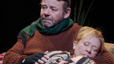 A man with glasses on his head and a green scarf sits on a sofa, comforting a woman in a patterned jumper and checked blanket who is resting against him, both appearing thoughtful or emotional.