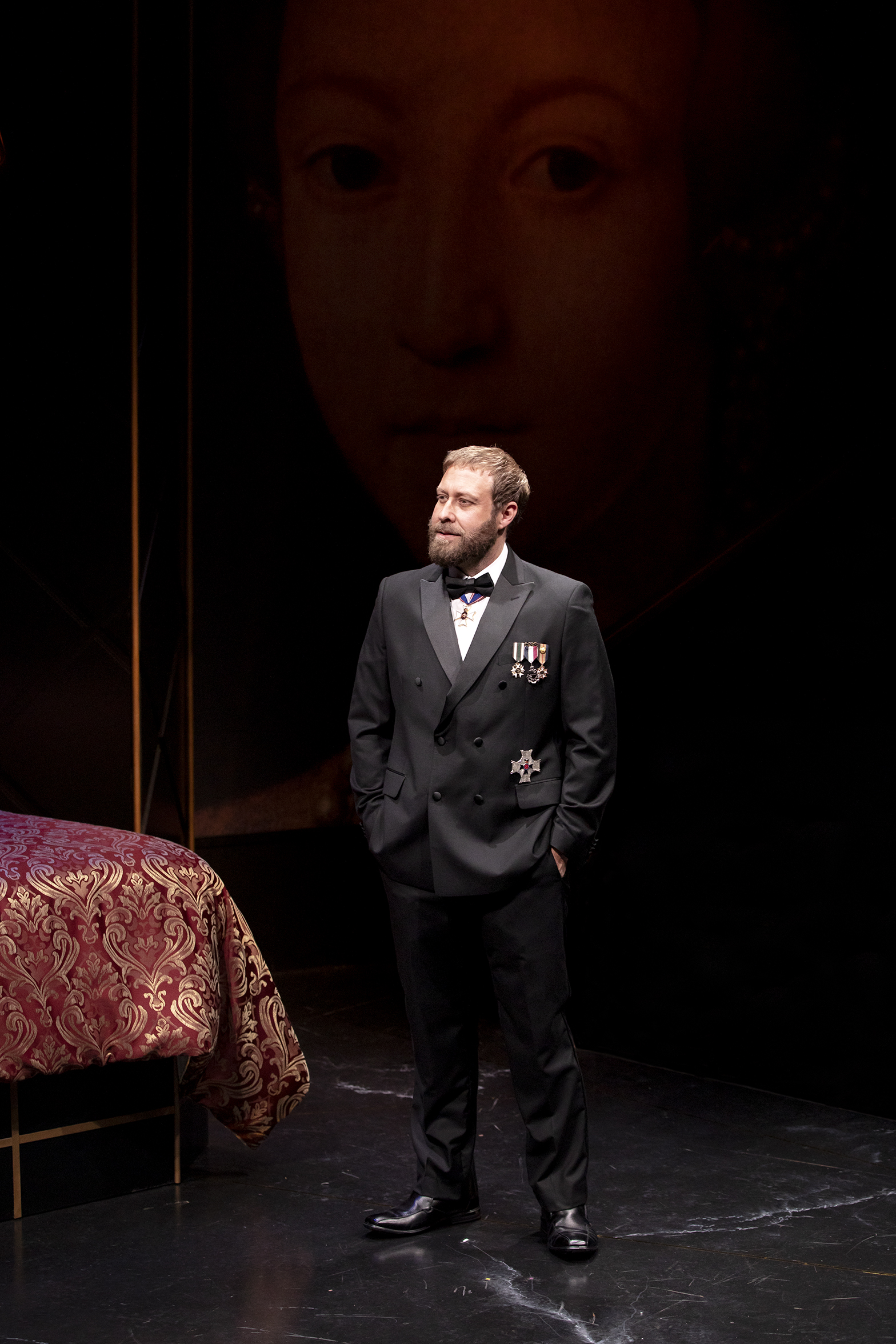 A man with a beard stands onstage in a black formal suit with medals, hands in his pockets. He stands beside a red and gold patterned bed, with a large portrait of a woman's face in the background.