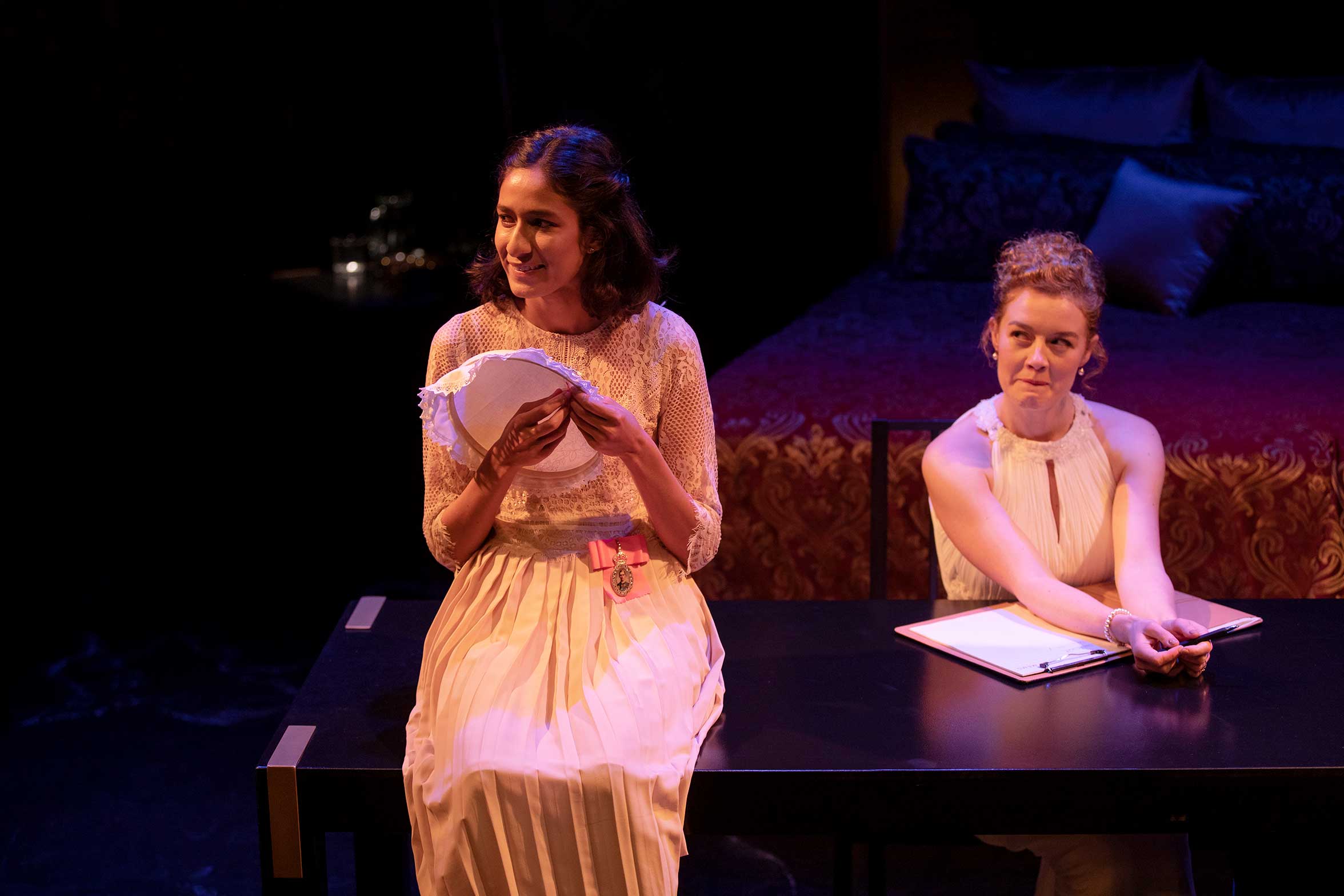 Two women in white dresses sit at a black table on stage; one holds papers and smiles, whilst the other sits with a notebook and pen, looking neutral. A bed with red and blue bedding is in the background.
