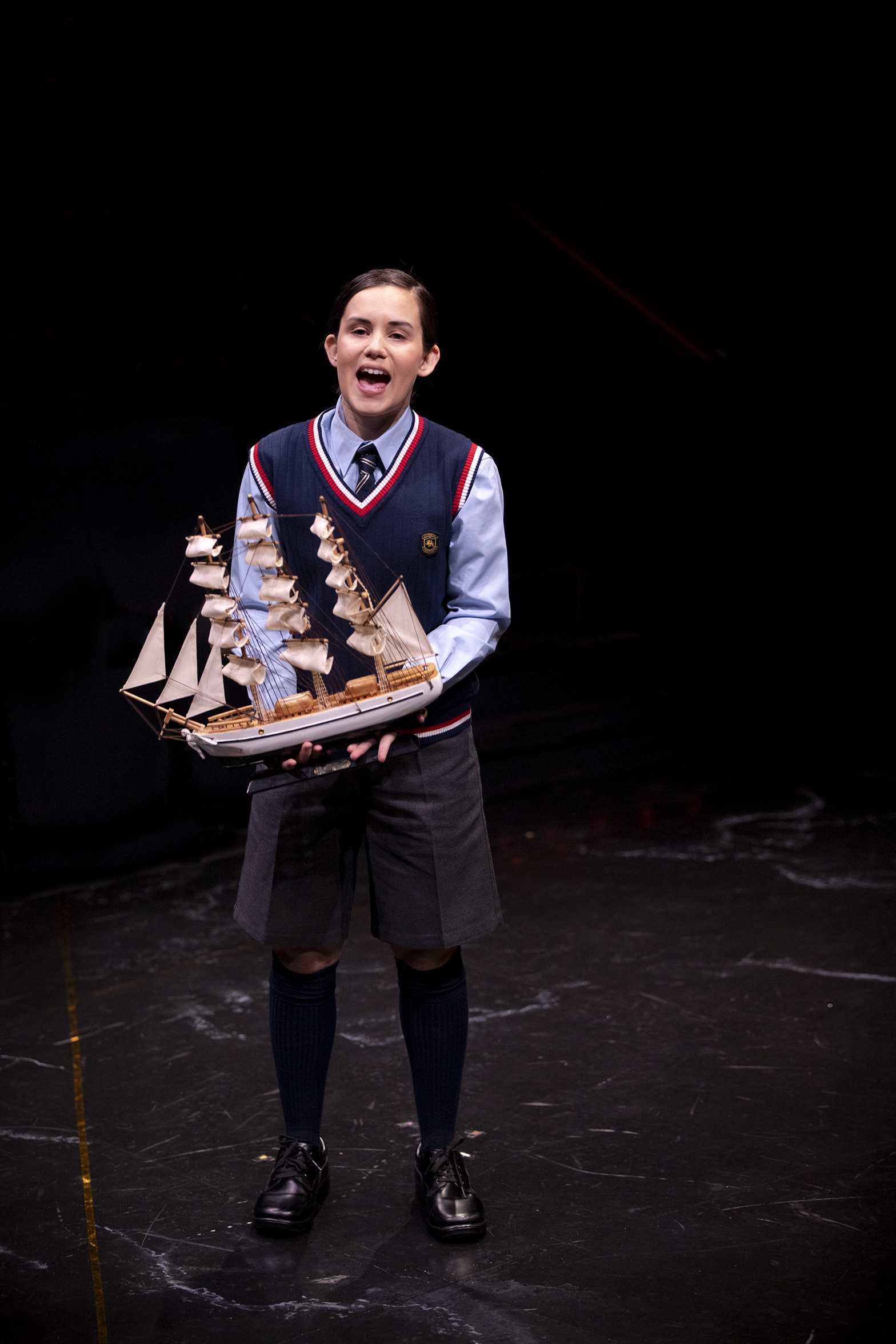 A pupil in a school uniform stands on a dark stage, holding a detailed model ship and speaking or singing, with an expressive look on their face.