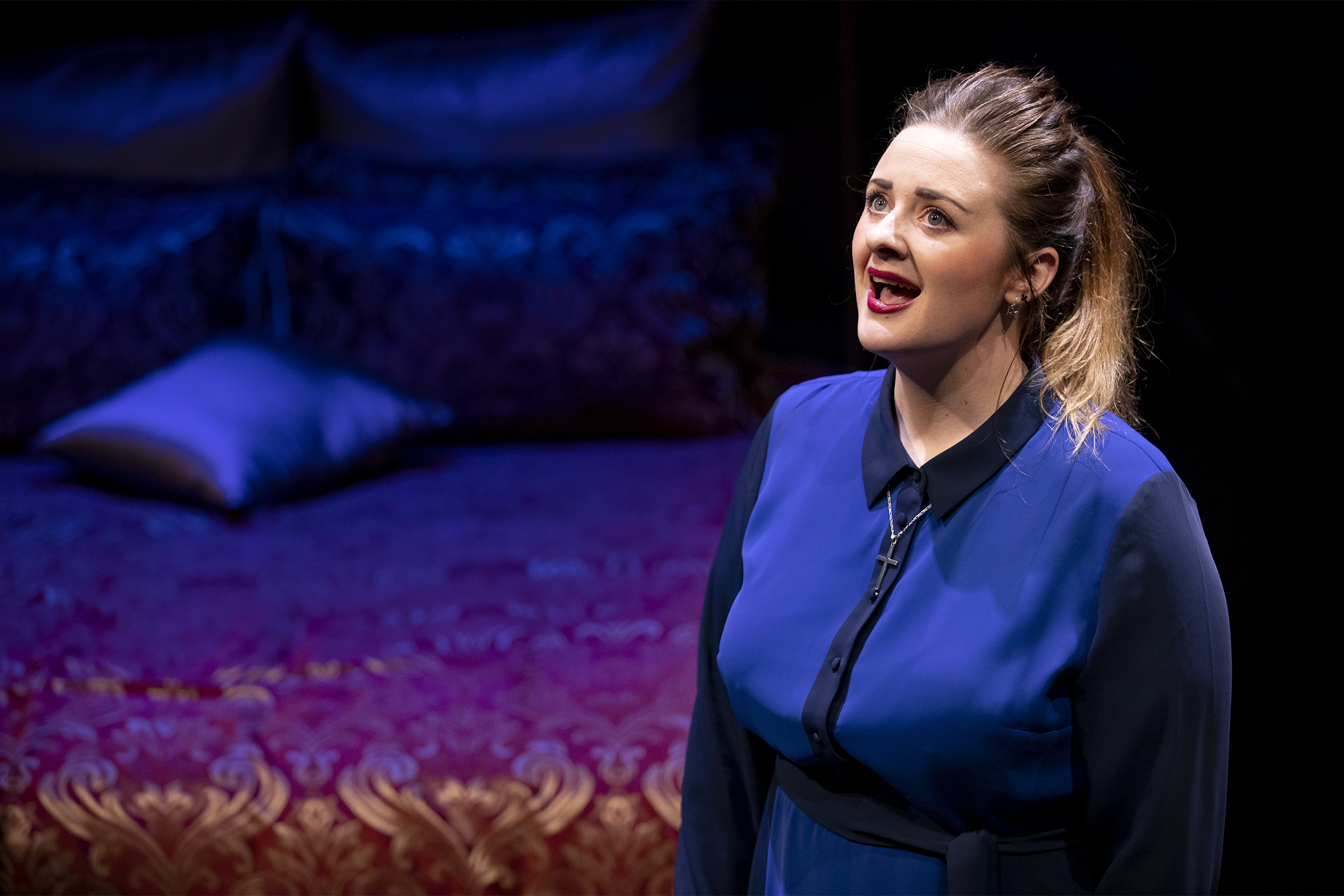 A woman in a blue dress sings passionately on stage, standing in front of a red and gold ornate bed with cushions, under dramatic lighting.