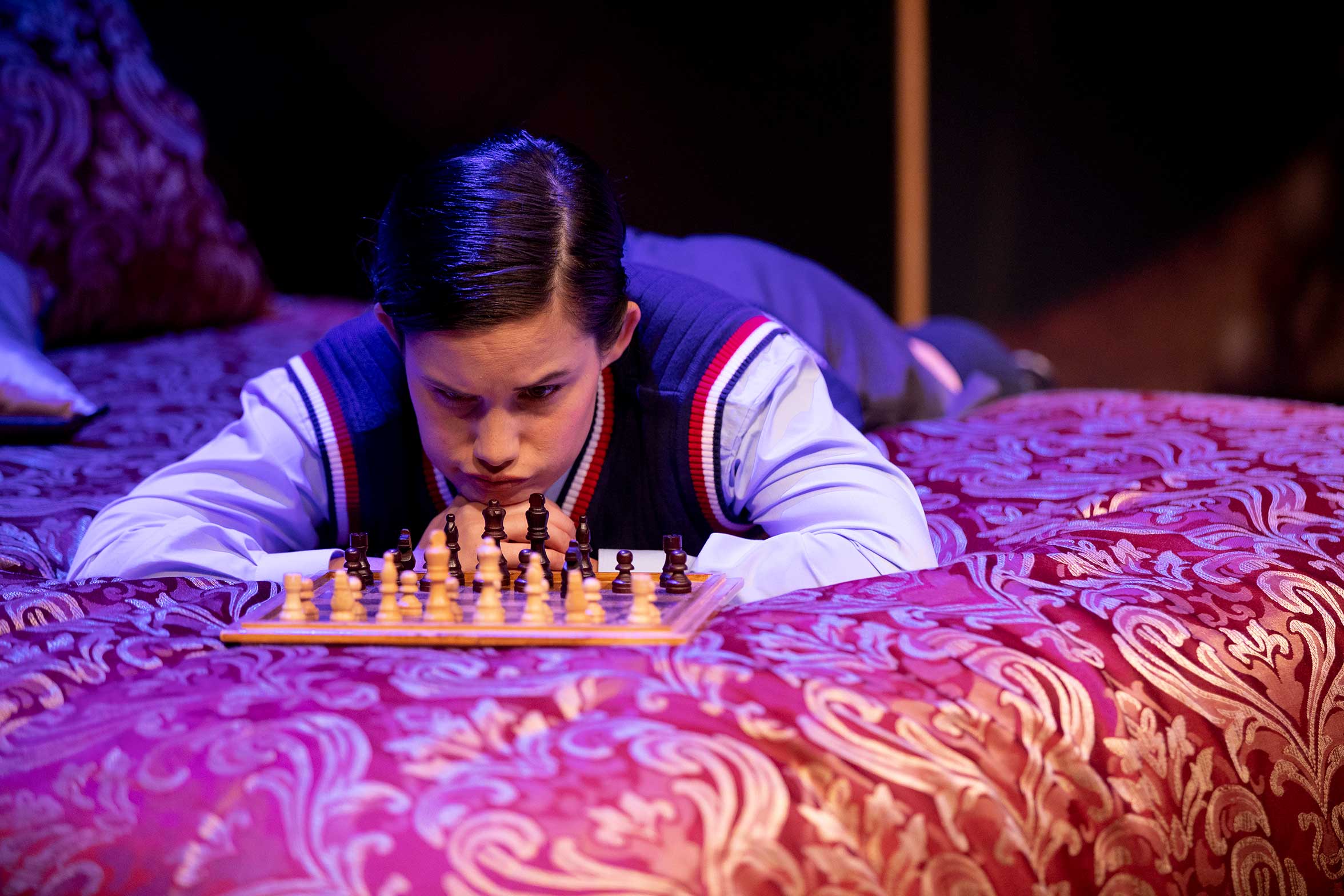 A young boy lies on a patterned bedspread, deeply focused on a chessboard in front of him, studying the pieces and contemplating his next move.