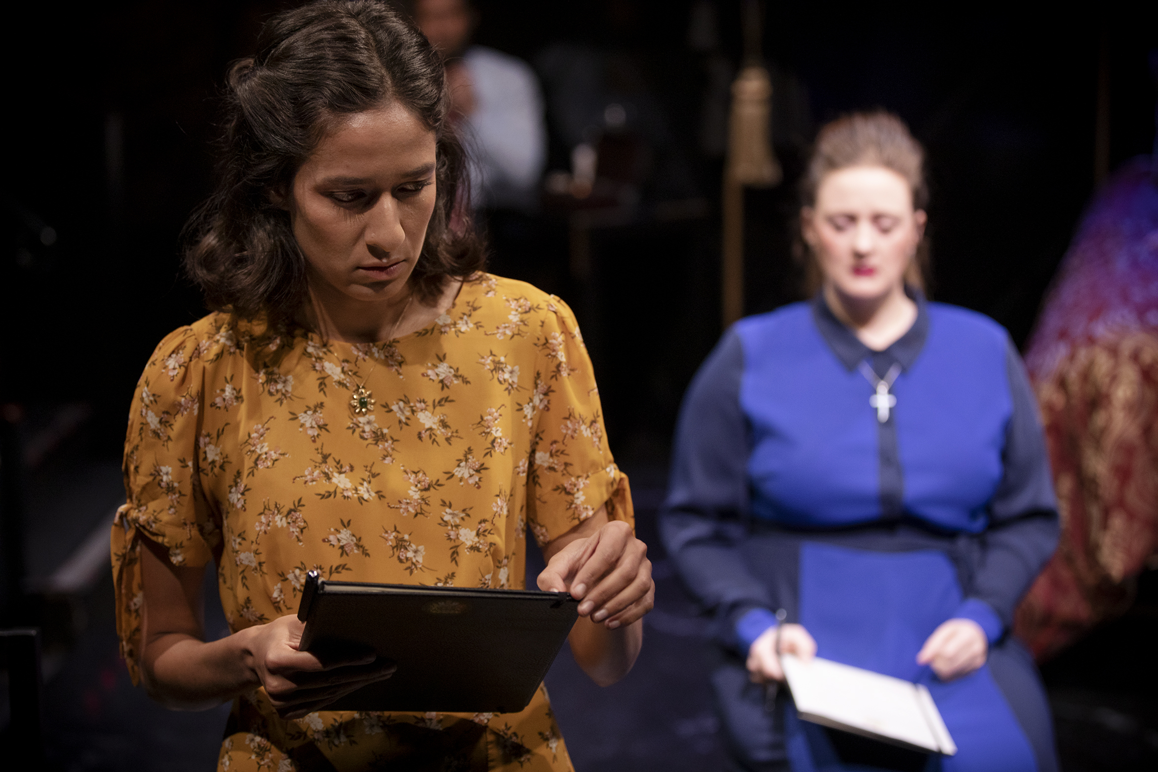 A woman in a yellow floral dress looks down at a tablet, whilst another woman in a blue dress with a cross necklace sits in the background holding papers. The scene appears to be from a stage performance.