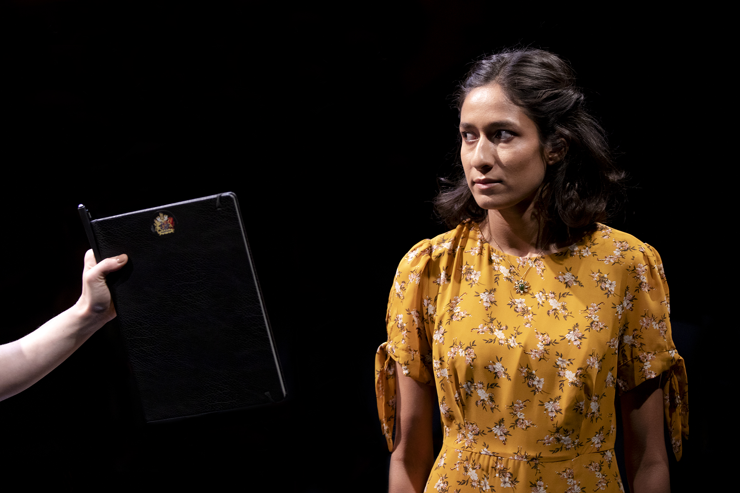 A woman in a yellow floral dress looks to the side with a serious expression as a hand holds a black folder towards her against a dark background.