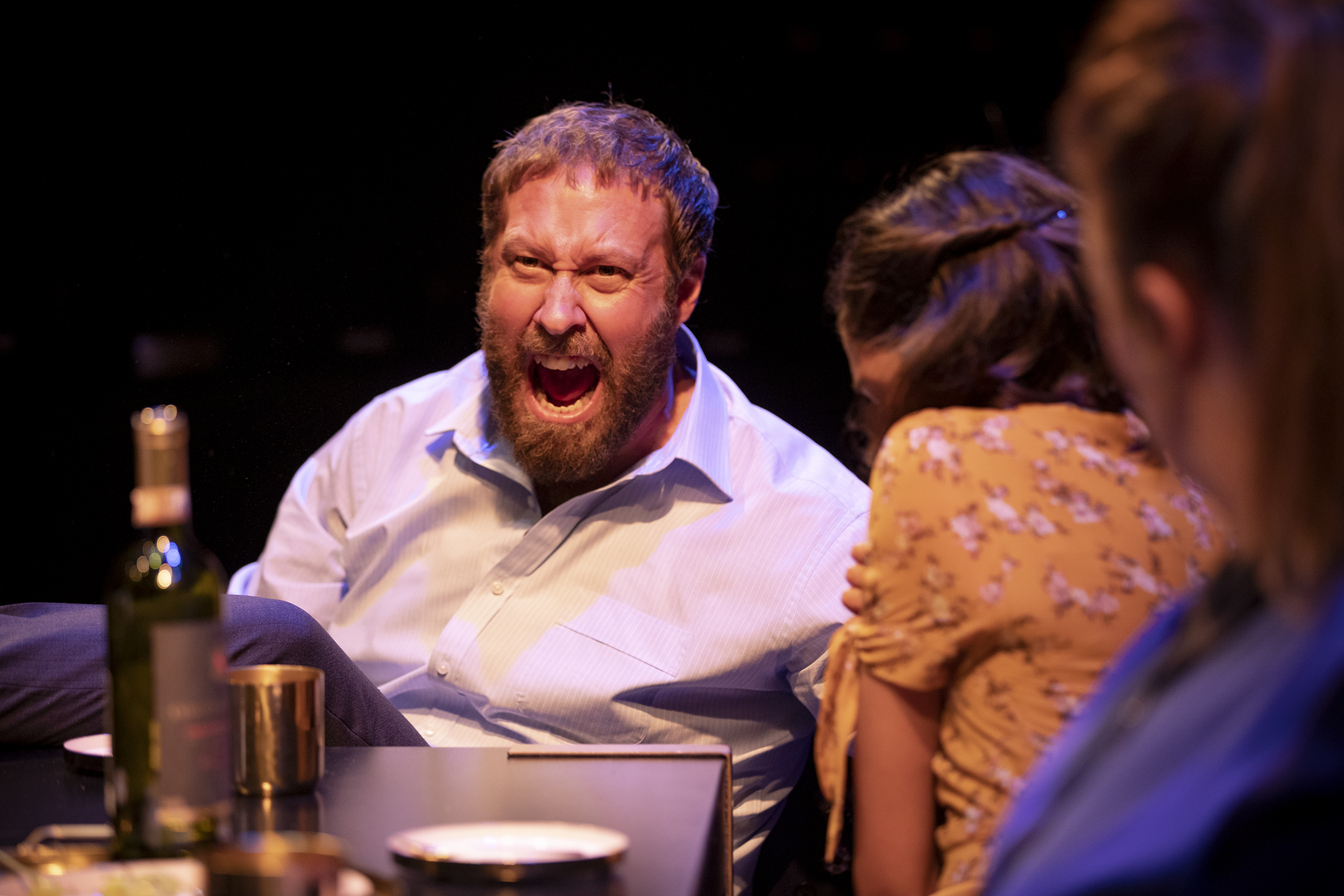 A bearded man in a dress shirt shouts passionately at a table, while a woman in a yellow dress next to him appears distressed and leans away; another person is partly visible in the foreground.