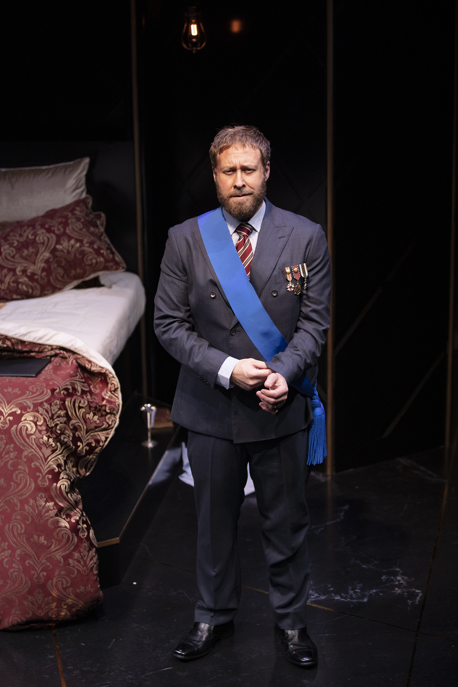 A man in a formal suit with medals and a blue sash stands on a dimly lit stage next to an ornate bed with red and gold bedclothes, looking serious and contemplative.