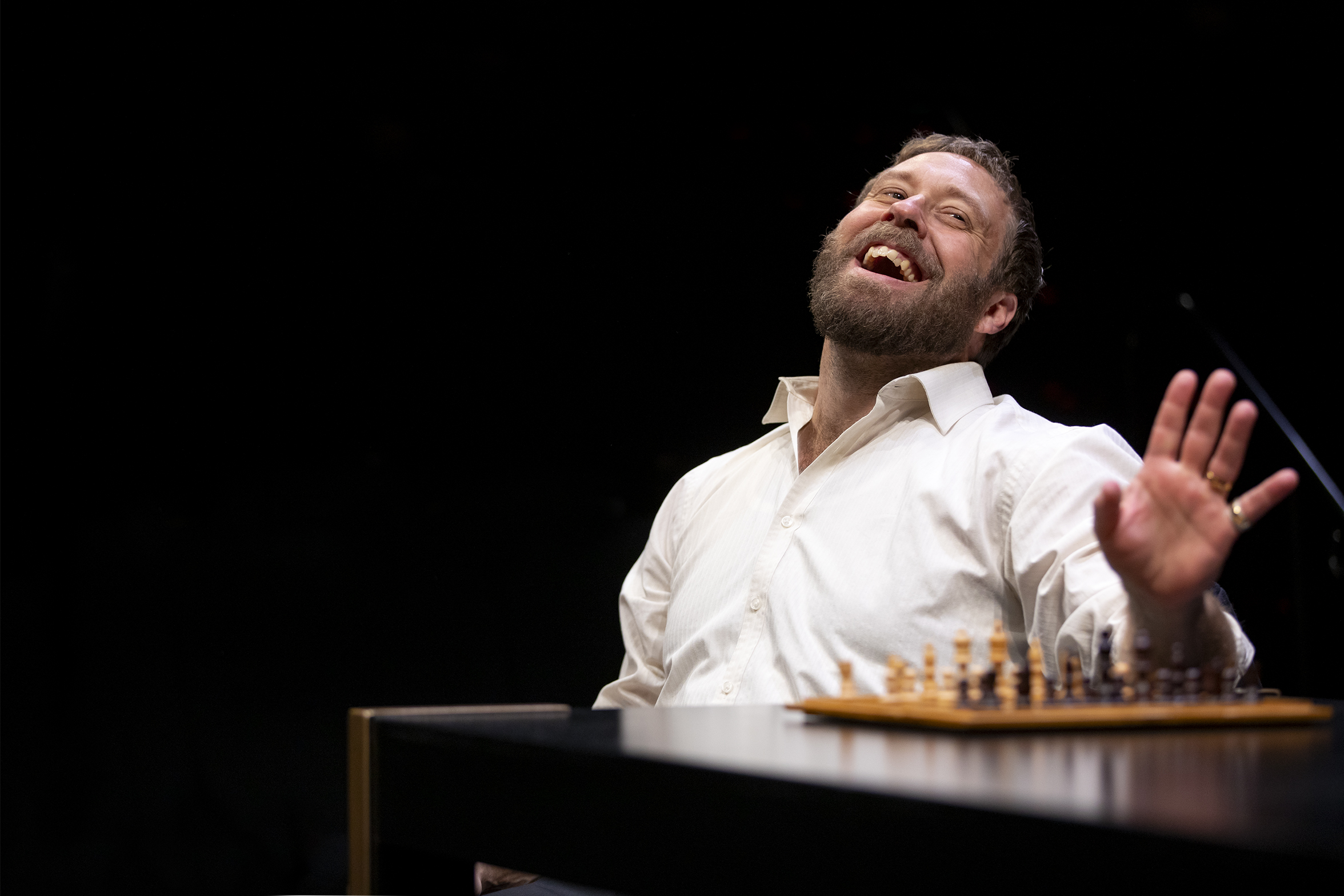 A bearded man in a white shirt sits at a table with a chessboard, leaning back and laughing with his hand raised, against a dark background.