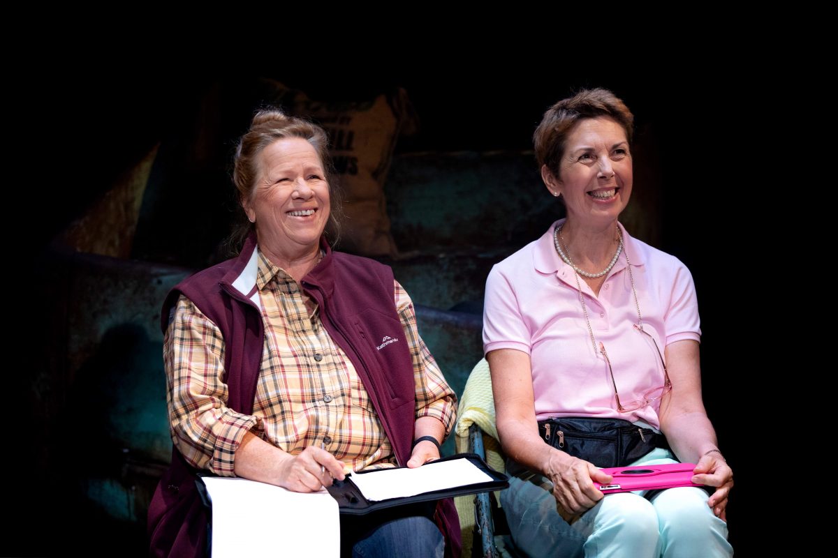 Two older women sit side by side, smiling and holding clipboards, appearing to be engaged in an activity or event. They are casually dressed and illuminated by stage lighting, with a dark background behind them.