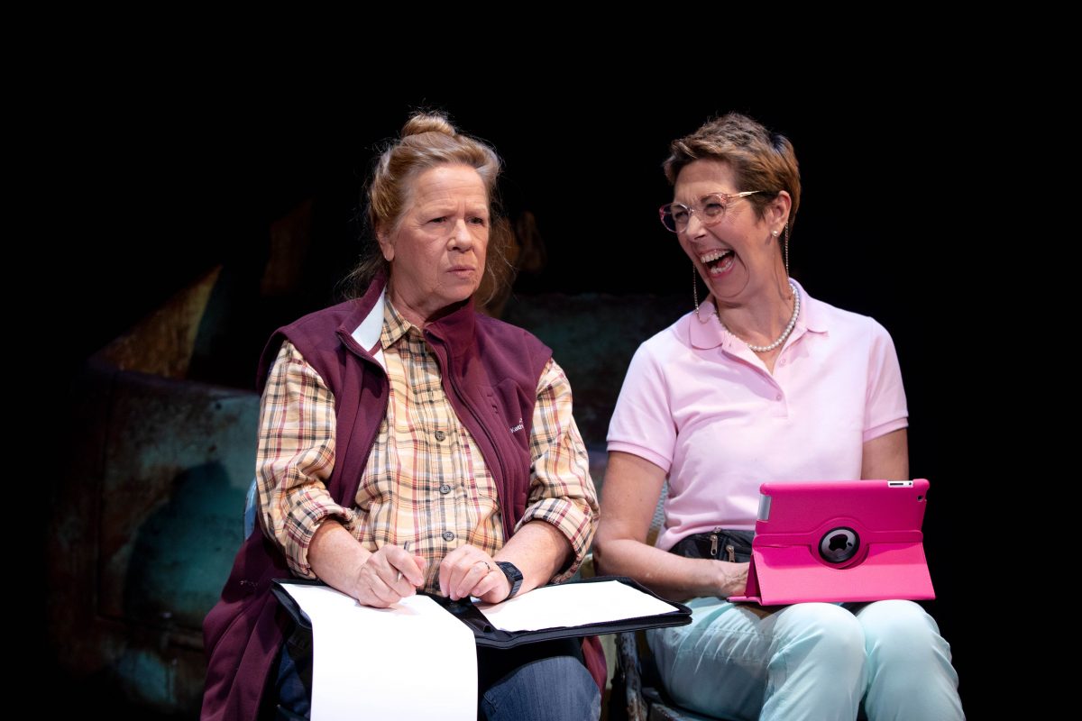 Two women sit side by side. The woman on the left looks serious, holding a pen and notepad. The woman on the right smiles and laughs, holding a bright pink tablet. Both appear to be engaged in a conversation or performance.