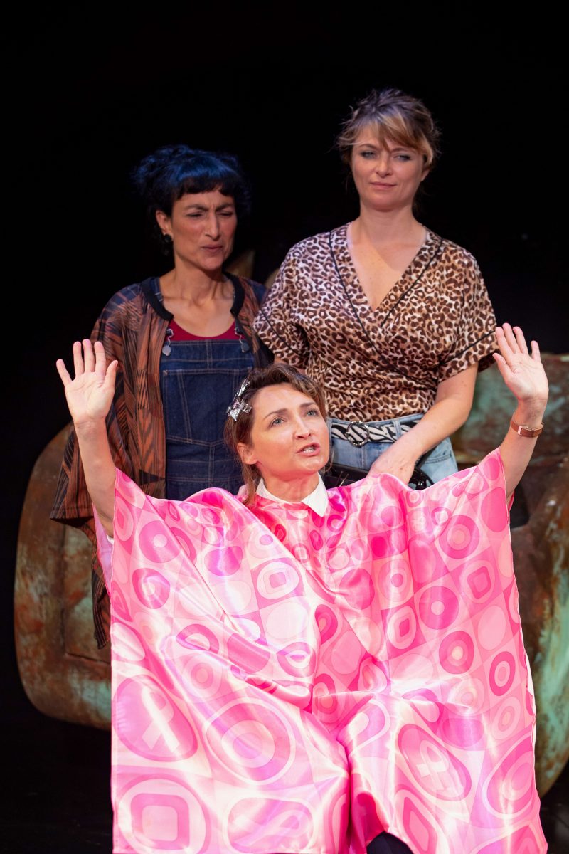 Three women are on stage; one is seated with her arms raised, covered in a pink geometric-patterned cape, whilst the other two stand behind her, looking on. The background is dark, highlighting the scene.