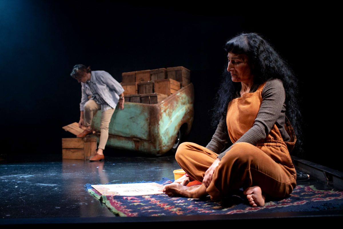 Two women on stage: one sits cross-legged on a patterned rug, looking thoughtful beside drawings; the other stands near a rusty car body, stacking wooden crates under dramatic lighting.