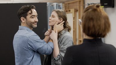A man gently holds a woman’s face as they rehearse a scene, while a third person observes them from behind. The three are in a room with wooden panels and rehearsal equipment.