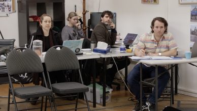 Four people sit at desks with laptops and papers in a casual office setting. Three of them are on the phone, and one is writing on paper. Empty chairs are in the foreground, and various office items are visible around them.