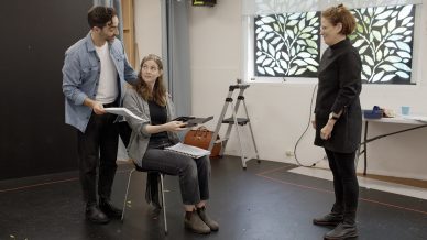 Three people in a rehearsal room: a man stands beside a woman who is sitting and holding scripts, whilst another woman stands opposite them, smiling. A ladder and a table are in the background.