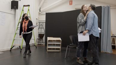 Three people in a rehearsal space: one person walks holding a script, wearing a mask; two others stand close together, one appearing to kiss the other's cheek. A green ladder and black set pieces are in the background.