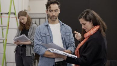 Three people stand indoors holding scripts. A woman in front gestures while talking to a man in a denim shirt; another woman in the background reads her script. A green ladder is partly visible on the left.