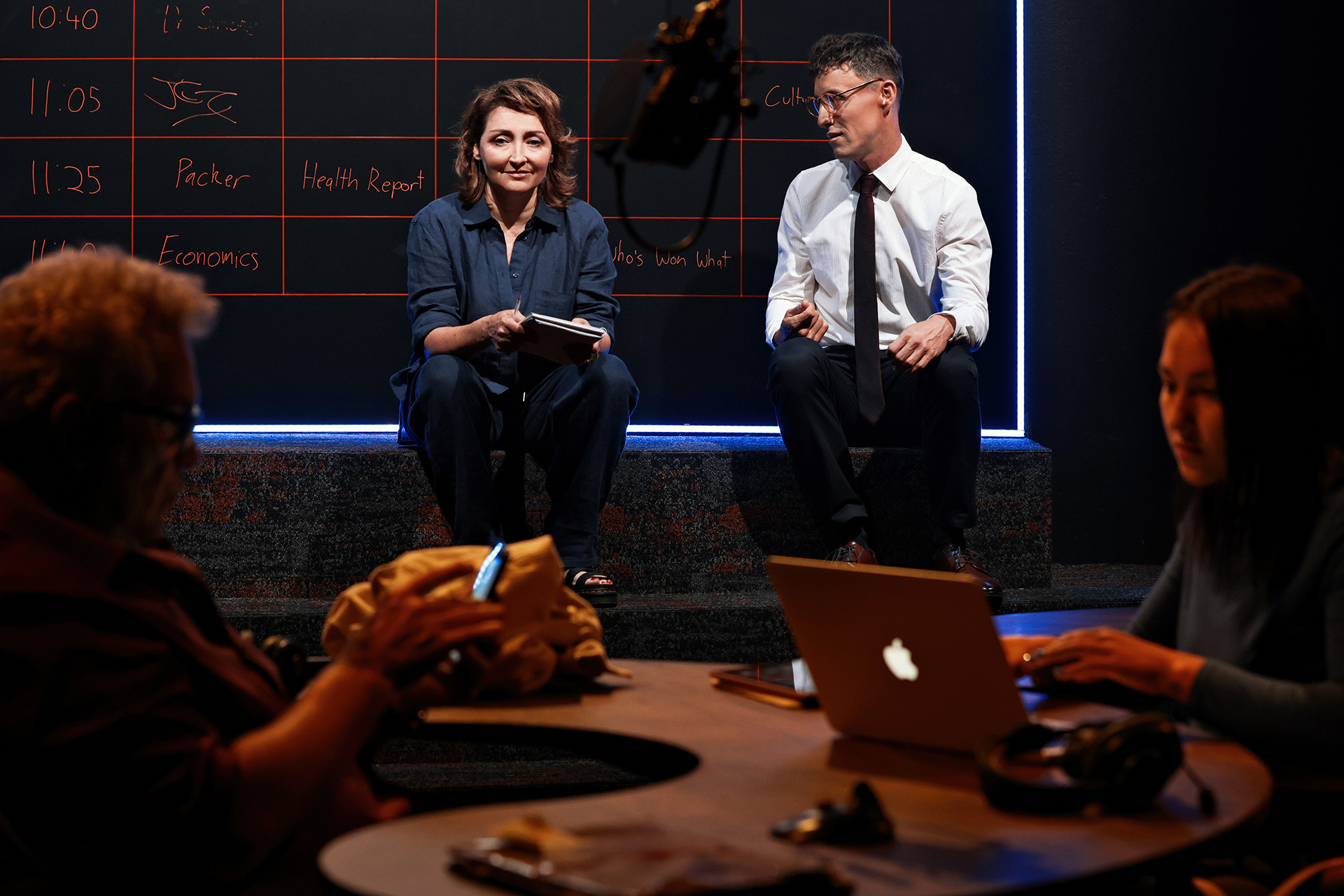 Four people are in a dimly lit room with a blackboard timetable. Two sit on a platform conversing, while two others work on laptops at a round table in the foreground. The atmosphere appears collaborative.