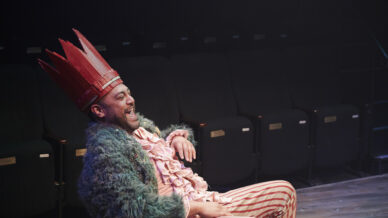 A person in a colourful costume with a red paper crown, frilly pink shirt, green jacket, and striped trousers laughs whilst sitting in a theatre with empty black seats in the background.