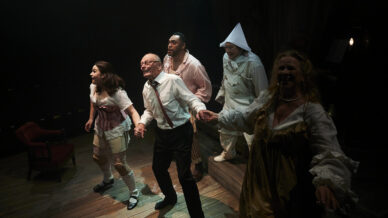 Five actors in colourful, vintage-style costumes hold hands and look outward on a dimly lit stage, appearing animated and expressive as they perform in a theatrical production.