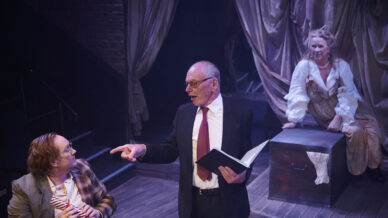 Three people perform on stage: one man in a suit reads from a book and points, another in a checked jacket and glasses holds a bow tie, and a woman in period costume sits on a chest in the background, draped curtains behind them.