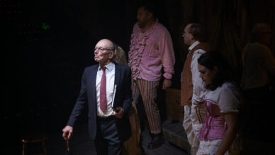 An older man in a suit stands on stage holding an umbrella, looking thoughtful. Behind him, four people in colourful, theatrical costumes watch intently in dim stage lighting.