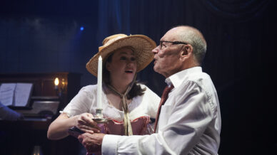 A young woman in a straw hat and period costume holds a candlestick beside an older man in glasses and a white shirt, who is looking ahead with concern. The background includes a piano and dim lighting.