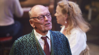 An older man in glasses, a patterned green dressing gown, white shirt, and red striped tie stands in focus, while a woman and another person in the background are out of focus. The scene appears to be indoors with warm lighting.