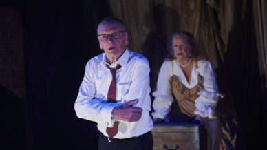 An older man in a white shirt and red tie stands with arms crossed, looking serious, while an older woman in period costume sits behind him on a chest against a dark, draped backdrop, both under stage lighting.