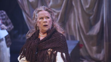 An older woman with long grey hair, wearing a brown shawl and white blouse, stands on stage with a surprised expression. The background features dramatic, draped curtains.