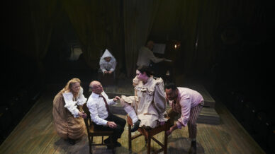 Five actors perform on a wooden stage; two are seated whilst others interact with them. One performer sits at a piano in the background. The scene is dramatic, with period costumes and dim, theatrical lighting.