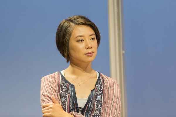 A young woman with short brown hair is standing with her arms folded, looking thoughtfully to the side. She is wearing a patterned blouse over a light top, against a plain blue background.