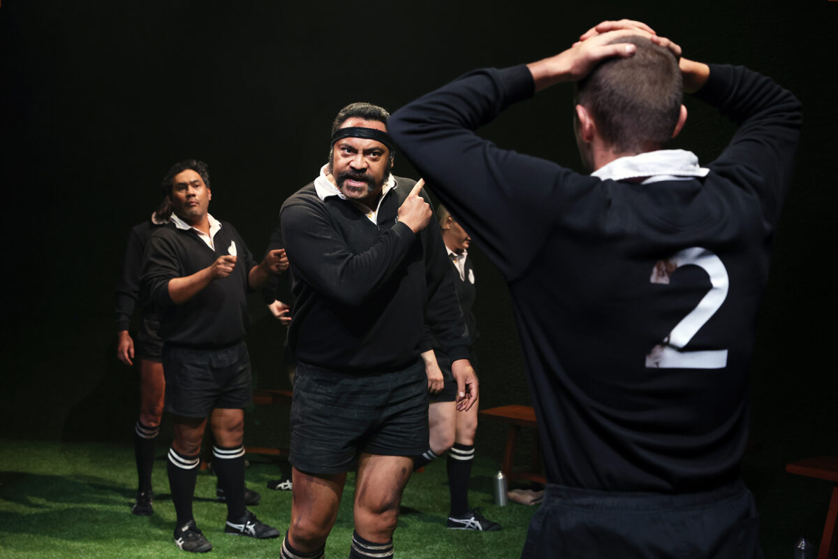 Four rugby players in black kits stand on a pitch; one man angrily points while another with 2 on his back holds his head, appearing frustrated. The scene is tense and dramatic, with other players in the background.