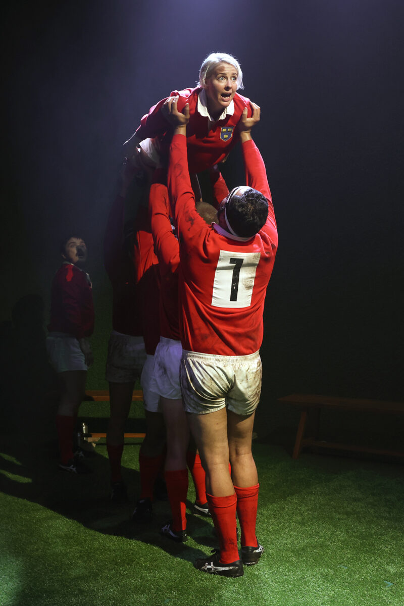 A rugby player wearing a red kit is lifted into the air by teammates during a line-out, with dramatic lighting highlighting the scene on a grassy pitch. The player looks ahead with a focused expression.