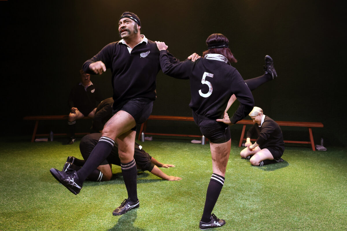 Four rugby players in black kits warm up on a grassy pitch; two stand at the front, raising their legs, while two others stretch on a bench in the background. The lighting is warm and theatrical.
