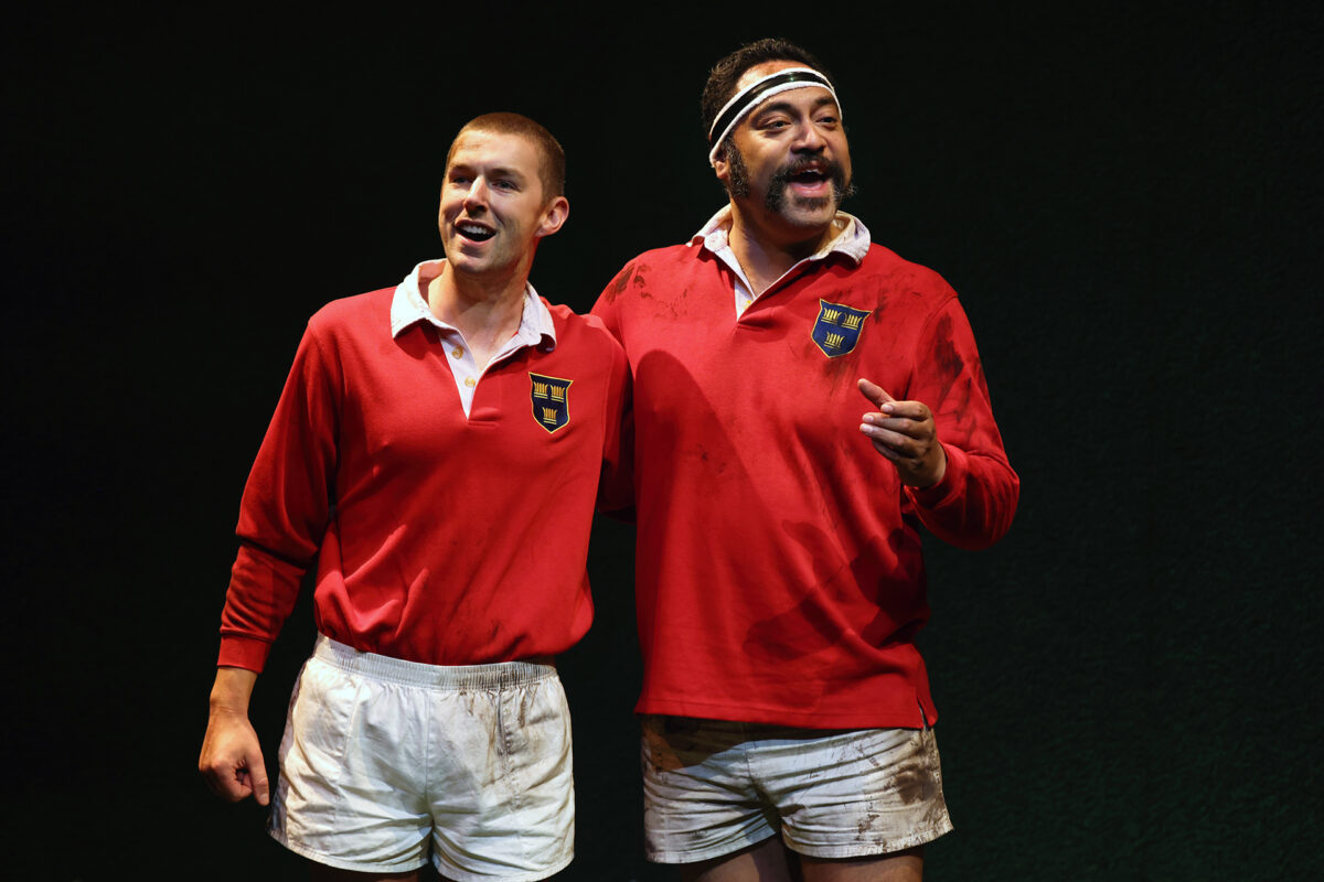 Two men wearing red rugby shirts and white shorts stand side by side, smiling. One has a headband and moustache, and both shirts have a shield logo. They appear on a dark, possibly stage-like background.