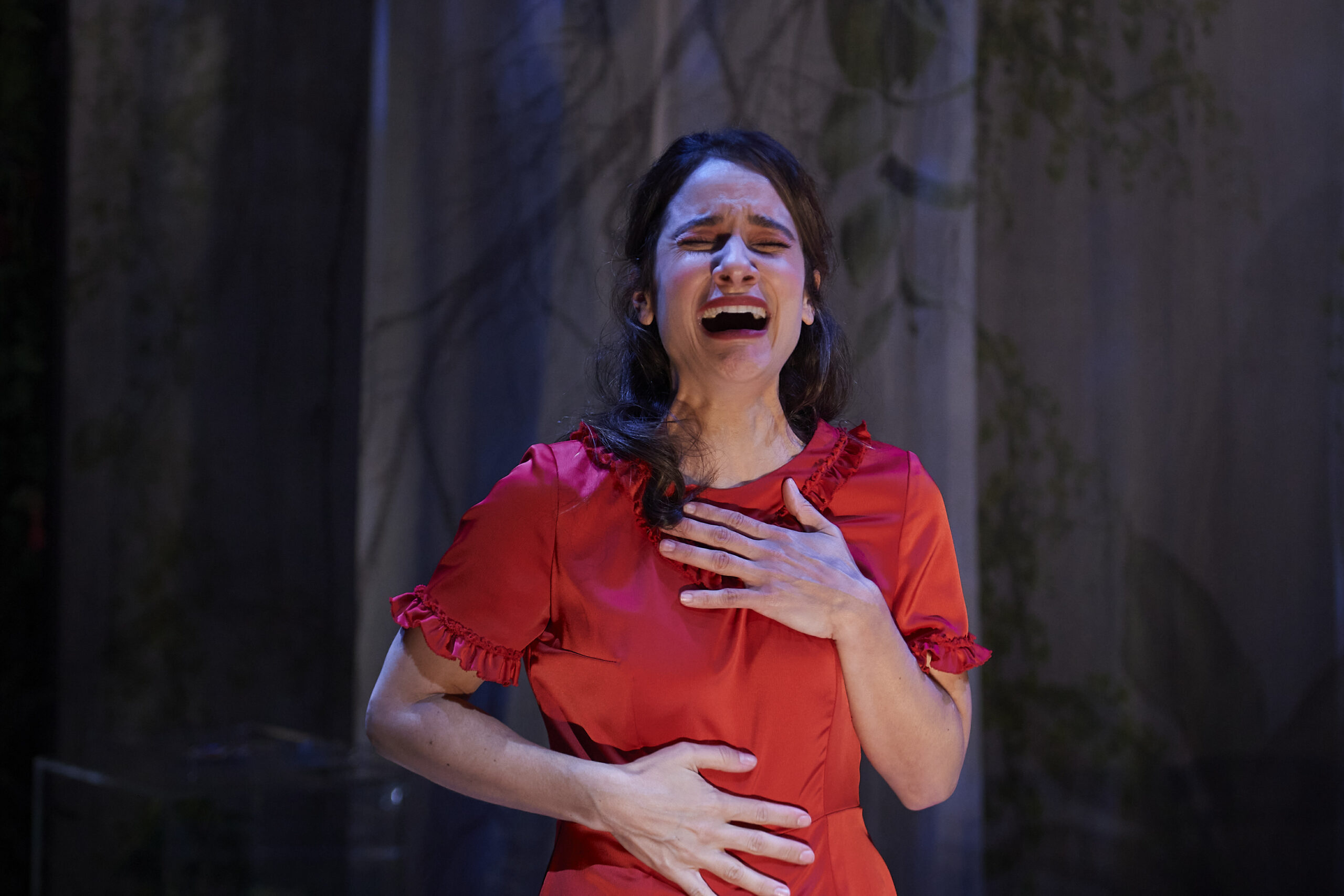 A woman in a red dress stands with one hand on her chest and the other on her stomach, eyes closed and mouth open in an emotional expression, as if singing or crying on stage.