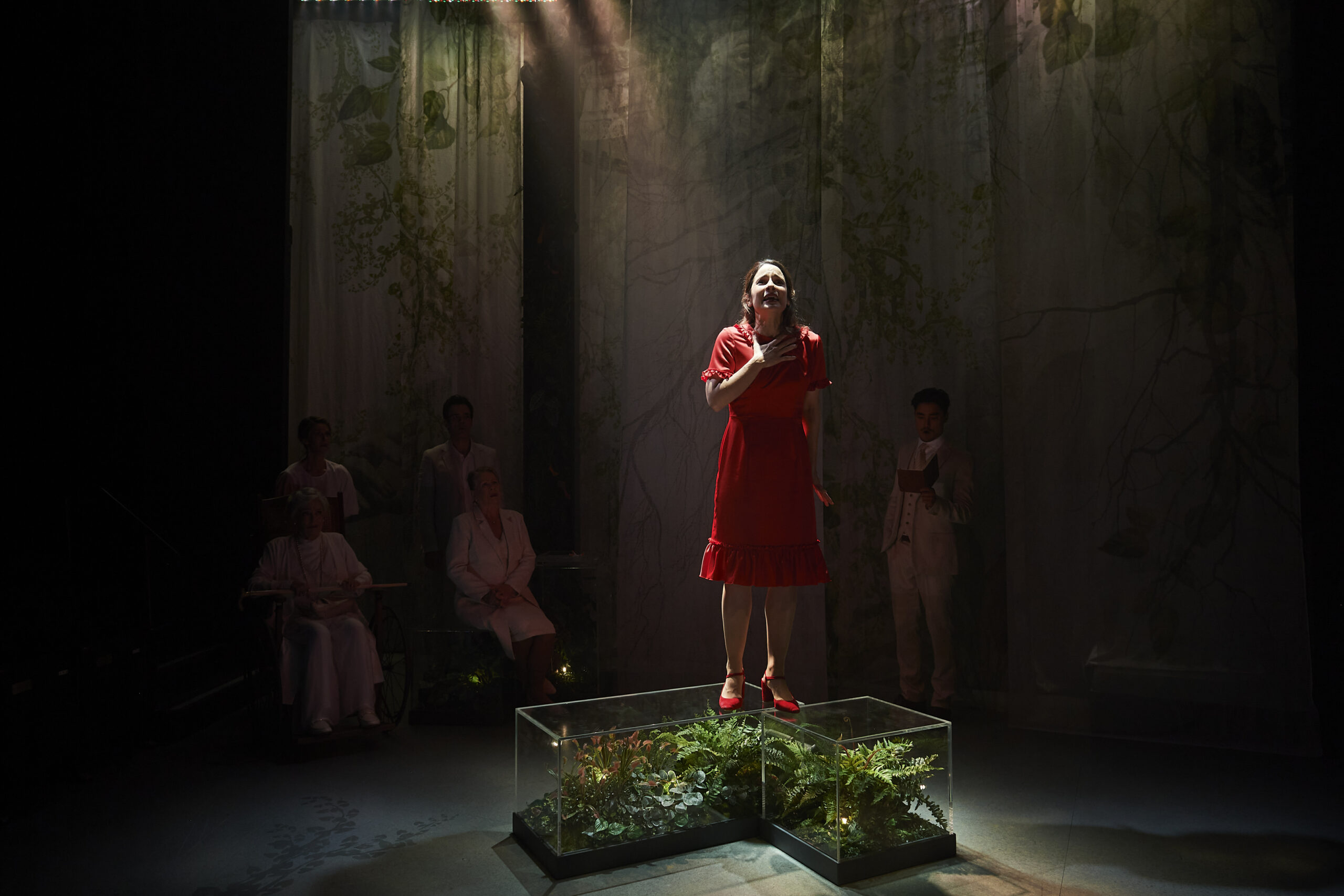 A woman in a red dress stands on a glass platform with green plants inside, centre stage under a spotlight. Four people in white or light clothing sit or stand in the dimly lit background, surrounded by leafy, nature-themed curtains.