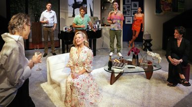 Seven adults are gathered in a modern living room, some standing and some seated. A woman in a floral dress sits on a chair, smiling, whilst others engage in conversation. The setting appears to be a social gathering.