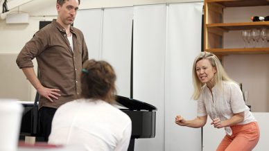 Three people are in a rehearsal room. A woman in white is acting with a dramatic expression and gesture, whilst a man in brown stands with his hand on his hip, looking serious. Another person sits with their back to the camera.