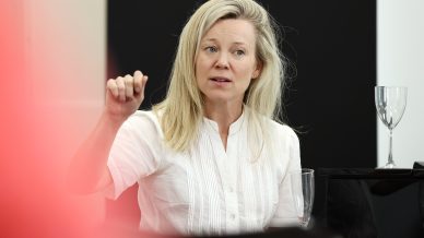 A woman with long blonde hair wearing a white blouse gestures with her hand whilst speaking. She sits near a wine glass, against a black and white background.