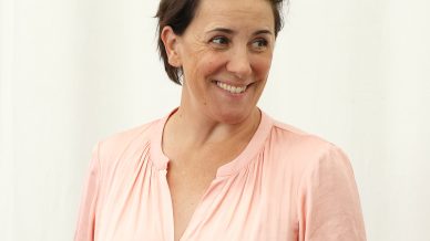 A person with short brown hair, wearing a light pink button-up shirt and a wristwatch, stands and smiles whilst gesturing with their right hand against a plain white background.