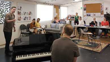 A man stands and speaks to a group of people seated in a creative studio, with a piano and a person sitting at it in the foreground. The group appears attentive, and mood boards and costume designs are visible on the walls.